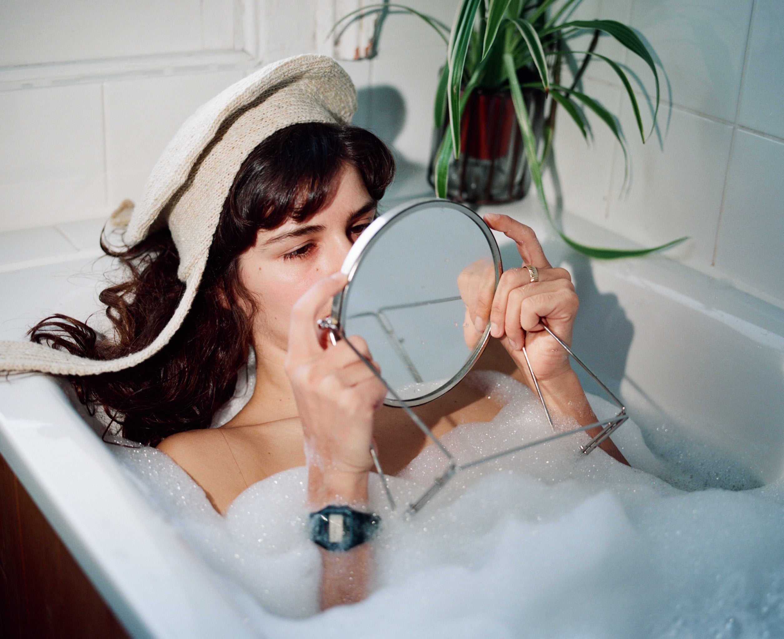 Model in a bubble bath looking at her reflection in a mirror wearing the classic hat in ecru